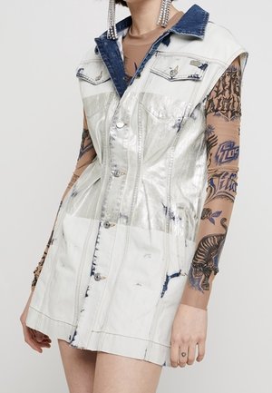 Kobieta ubrana w bezrękawnikową białą sukienkę dżinsową o srebrzystym połysku z niebieskim kołnierzem, na przezroczystej bluzce z długim rękawem z nadrukiem tatuażu oraz dużymi wiszącymi kolczykami.