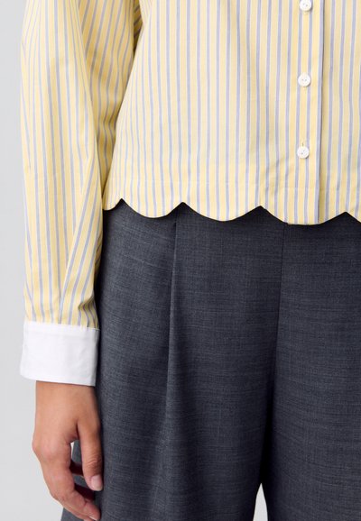 Blusa a righe gialle e blu con orlo sagomato, abbinata a pantaloni grigi. Presenta un polsino bianco e bottoni lungo la parte anteriore.