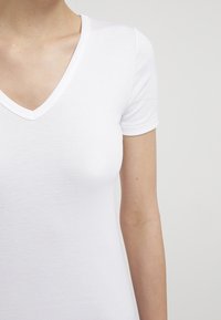 Vit kortärmad V-ringad t-shirt av ribbat bomullstyg; slät textur med figurnära design, visar en närbild av halsringningen och ärmen.