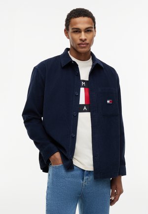 Noor mees, kes kannab sinist nööpidega särki valge logoga T-särgi peal, üks käsi taskus, seisab tavalise valge tausta ees.