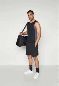 Mann in schwarzem ärmellosem Oberteil und Shorts, der eine schwarze Sporttasche über der Schulter hält und vor einem einfarbigen hellgrauen Hintergrund steht.