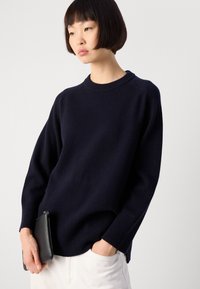 Nainen, jolla on lyhyet mustat hiukset, pukeutuneena tummansiniseen neuleeseen ja valkoisiin housuihin, pitää yhtä kättään taskussa ja toisessa on pieni musta clutch.