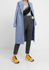 Lichtblauwe lange jas, grijze getailleerde top, donkergrijze slim fit broek en felgele sneakers met zwarte accenten en ritsen.