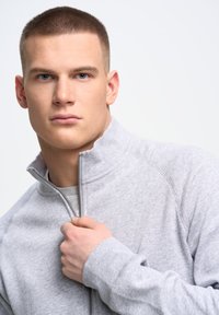 Szary rozpinany sweatshirt wykonany z miękkiego, teksturowanego materiału, z wysokim kołnierzem i ściągaczami przy mankietach. Model trzyma suwak jedną ręką.