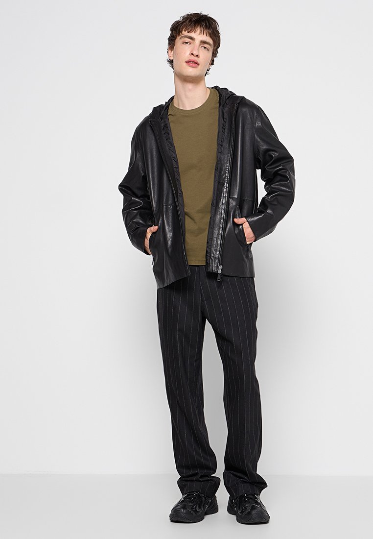Junger Mann steht mit den Händen in den Taschen, trägt eine schwarze Lederjacke, ein olivgrünes T-Shirt, schwarze Nadelstreifen-Hosen und schwarze Sneaker.