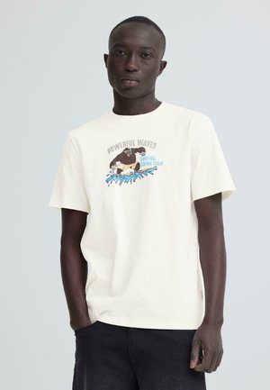 Ung mand iført en hvid T-shirt med en grafisk fremstilling af en gorilla, der surfer, og teksten "Powerful Waves Surfing Diving Team," stående med den ene hånd i lommen.