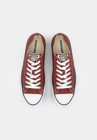 Rde platnene superge z belimi vezalkami, belimi gumijastimi kapicami in črnimi detajli ter klasičnim nizkim dizajnom. Opremljene z blagovno znamko "Converse All Star".