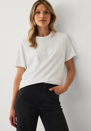 Kvinde med lyst brunt hår iført hvid T-shirt med blomsterapplikationer og sort højtsiddende jeans, stående op ad en hvid væg.