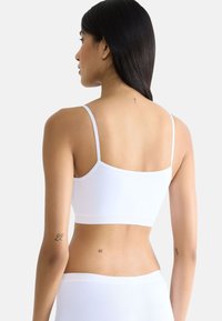 Witte bralette met dunne bandjes, gladde textuur en een eenvoudig rechthoekig ontwerp, gezien vanaf de achterkant. Tattoos zichtbaar op de onderarm en rug.
