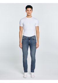 Fiatal férfi egyenes tartással, sima fehér pólót, kék farmert és fehér tornacipőt viselve, világosszürke háttér előtt.