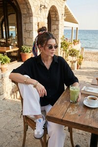 Femme portant des lunettes de soleil et un foulard coloré, assise à une table d’un café en bord de mer avec des boissons et un livre, bâtiment en pierre et océan en arrière-plan.