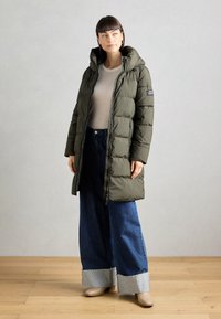 Olijfgroene pufferjas met een capuchon, ritssluiting en patchdetails, gedragen over een lichtgrijze top en een wijde, donkerdenim spijkerbroek met omslagen.
