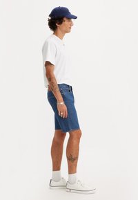 Blue denim shorts, white cotton t-shirt, and white sneakers with navy accents. Model has curly hair and visible tattoos on arms.