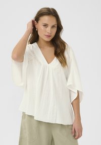 Blusa bianca leggera con scollo a V, maniche ampie e delicate decorazioni ricamate. Tessuto testurizzato con pieghe sulla parte frontale.