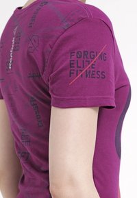 Fuchsiové tričko s krátkým rukávem vyrobené z bavlny. Obsahuje potištěný text: "FORGING ELITE FITNESS" a "CrossFit" v černé barvě. Jednoduchý design s kulatým výstřihem.