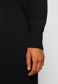 Main droite détendue sur le côté, portant un vêtement en maille noire à manches longues avec un poignet ajusté sur un fond uni.