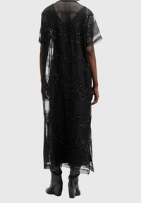 Zwarte doorzichtige jurk met horizontale strepen van pailletten, losse pasvorm, korte mouwen en een lange zoom. Draagt over strakke zwarte pantalons en laarzen.