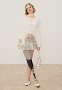 Νέα γυναίκα φορώντας ανοιχτόχρωμο ζακέτα, φούστα με σχέδια και τεχνητό πόδι κάτω από το γόνατο, κρατάει μια ανοιχτό ροζ τσάντα.