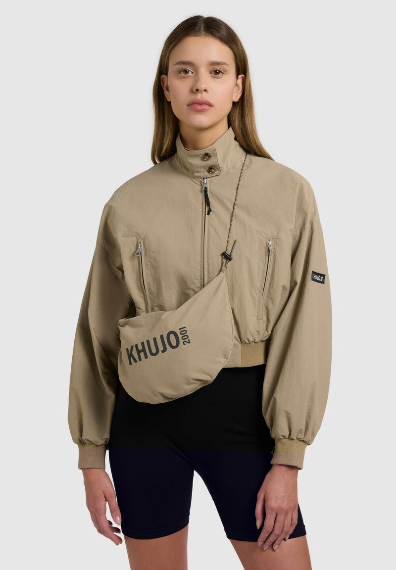 Femme portant une veste beige à fermeture éclair avec un sac bandoulière assorti portant l'inscription « KHUJO 2001 » et un short de cyclisme noir, debout devant un fond neutre.