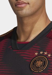 Musta jalkapallopaita, jossa on punainen ja kultainen abstrakti kuvio, V-pääntie, pyöreä brodeerattu logo ja neljä tähteä sen yläpuolella.