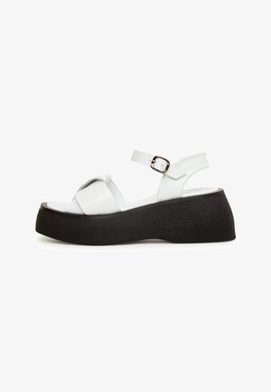 Weiße Plattform-Sandalen mit einer breiten, strukturierten schwarzen Sohle, verstellbarem Knöchelriemen und überkreuztem Oberteil-Design. Glatte lederähnliche Oberfläche.