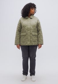 Πρασινοελαιώδες καπιτονέ μπουφάν με μεγάλες μπροστινές τσέπες, στρογγυλή λαιμόκοψη και μακριά μανίκια. Φοράται πάνω από γκρι τζιν και λευκά αθλητικά παπούτσια.