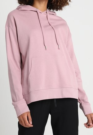 Persoon draagt een lichtroze oversized hoodie met een voorvak en trekkoorden, gecombineerd met zwarte broek tegen een effen achtergrond.