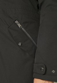 Veste noire avec une poche zippée, un bouton-pression et une texture lisse. Le design comprend un col montant et une coupe ajustée.