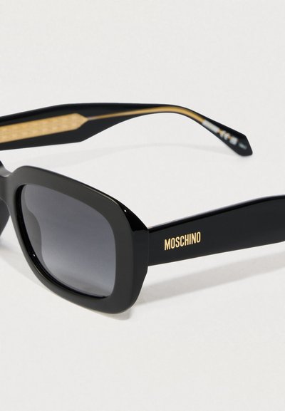 Melni taisnstūra saulesbrilles ar tumšiem stikliem un zeltītu "Moschino" logo uz kājas, fotografētas uz baltas fona.