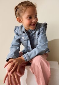 Giovane ragazza con i capelli intrecciati che indossa una giacca di jeans blu e pantaloni rosa, seduta, sorridente e guarda verso destra.