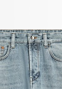 Lichtblauwe spijkerbroek in close-up met zicht op voorste knoop, rits, riemlussen en zakken met zichtbare stiksel details.