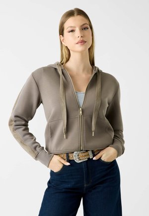 Jeune femme portant un sweat à capuche taupe zippé avec de larges cordons, ceinture marron décorée, et jean bleu foncé, debout les mains dans les poches.