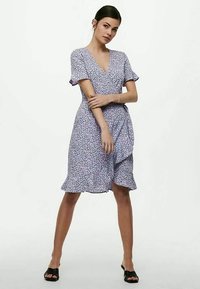 Femme debout dans une robe enveloppante à manches courtes de couleur lilas clair avec un ourlet à volants, portant des talons ouverts noirs sur un fond blanc uni.