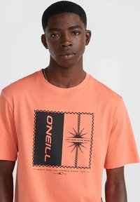 Camiseta de algodón de manga corta en color coral, con un estampado gráfico negro que incluye texto y formas geométricas centradas en el pecho.