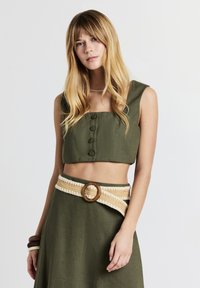 Donna con lunghi capelli biondi che indossa un crop top senza maniche color verde oliva e una gonna alta coordinata con una cintura intrecciata con fibbia di legno.