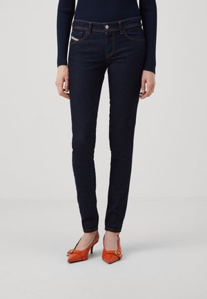 Femme portant un jean slim bleu foncé et des escarpins pointus orange vif avec un détail boucle sur un fond uni clair.