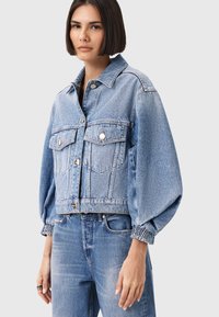 Giacca in denim azzurro chiaro, cropped, con maniche lunghe a sbuffo, due tasche sul petto e dettagli in bottone argentato. Tessuto liscio, vestibilità comoda.