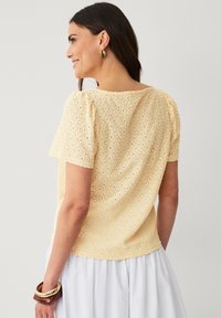 Lys gul bluse med et struktureret, broderet design med rund halsudskæring og korte puffærmer. Båret sammen med en hvid nederdel.
