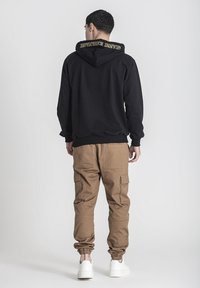 Sudadera negra con capucha y logo en la parte posterior, combinada con pantalones cargo marrones con puños elásticos y bolsillos laterales. Zapatillas blancas.