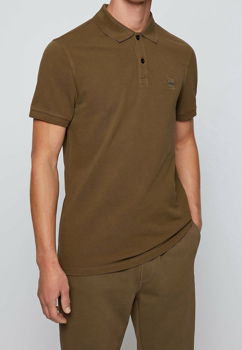 Homme portant un polo marron à manches courtes avec deux boutons noirs et un petit logo sur la poitrine gauche, assorti à un pantalon marron.
