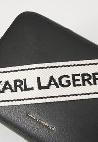 Svart lærpung med et bredt hvitt bånd med "KARL LAGERFELD" i fet svart skrift, supplert med et gulllogo-aksent.