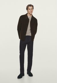 Giacca button-up marrone scuro sopra un maglione grigio chiaro, abbinata a pantaloni scuri e scarpe nere. Il tessuto appare liscio con una vestibilità strutturata.