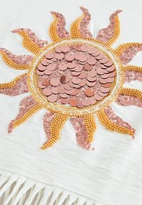 Vit tyg med en soldesign som är gjord av rosa och guld paljetter, med orange pärlaccenter och frans längs den undre kanten.