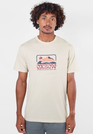Homme aux cheveux bouclés portant un T-shirt beige clair avec un graphisme coloré de montagne et de soleil et le texte "OXBOW" sur le devant.