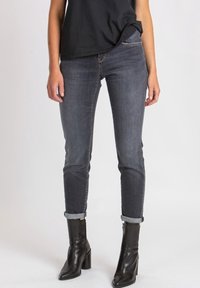 Mujer vestida con jeans oscuros ajustados y remangados, botas de tacón negras hasta el tobillo y una blusa negra, contra un fondo claro y liso.