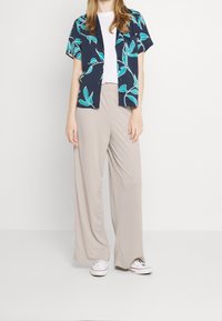 Koszula z krótkim rękawem w kolorze granatowym z turkusowym wzorem liści, zestawiona z luźnymi beżowymi spodniami. Pod spodem biała koszulka i białe trampki.