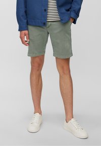 Homme portant un short vert, des sneakers blanches, une chemise rayée et une veste bleue, avec une main dans sa poche, devant un fond uni.