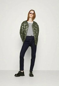 Casaco bomber acolchoado verde, camisa às riscas pretas e brancas, jeans escuros e sapatos pretos robustos. Óculos de sol completam o conjunto.