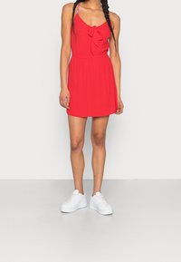 Nainen, jolla on päällään ilman hihoja punainen mekko, jossa on edessä solmu, ja valkoiset platform-nilkkurit, seisoo yksinkertaista valkoista taustaa vasten.