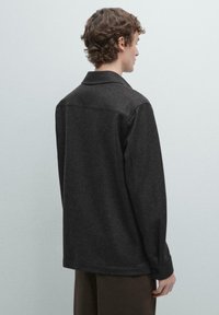 Ciemnoszara wełniana kurtka o luźnym kroju, z zaokrąglonym kołnierzem i długimi rękawami zakończonymi mankietami na guziki, widziana od tyłu.